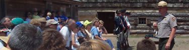 a park ranger stands in the middle of a crowd speaking on the fort parade ground. the summer sun is his spotlight