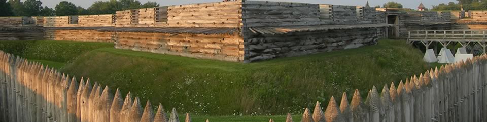 large wooden spikes jut out from a large wooden angular wall lit by sunlight. verdant grass surrounds it.