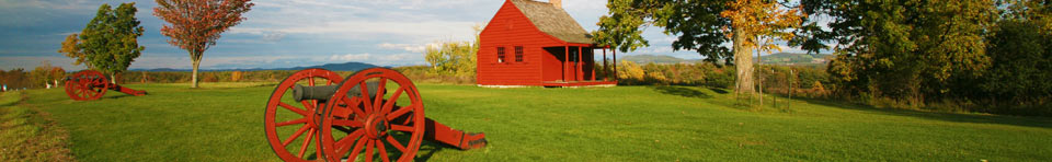 Scenic rural landscape with green lawn, cloud-flecked blue sky, and autumn foliage punctuated by a small, deep red farmhouse and red cannon carriages.