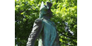 A green statue stands among trees, it has a soldiers uniform on, with a cocked hat, and a sword. A green statue stands among trees, it has a soldiers uniform on, with a cocked hat, and a sword.