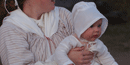 A small baby is held in its mother's arms. It is wearing a white cap, and a white fabric sheet as a covering. Its mother clutches it tightly A small baby is held in its mother's arms. It is wearing a white cap, and a white fabric sheet as a covering. Its mother clutches it tightly