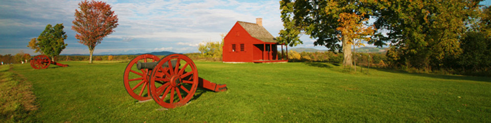 Scenic rural landscape with green lawn, cloud-flecked blue sky, and autumn foliage punctuated by a small, deep red farmhouse and red cannon carriages.