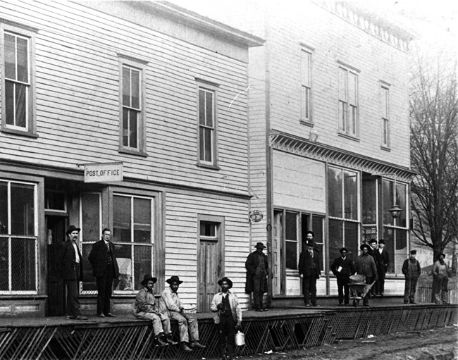 Historic image of two buildings with a sign saying Post Office in front of one and men on the porches