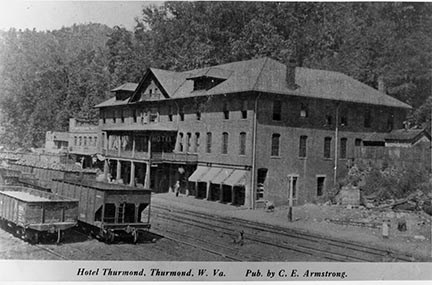 A large four story building with a veranda out front right next to several sets of railroad tracks with freight cars on them