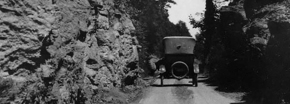 An old fashion turn of the century era car driving on a dirt road next to a rock face.