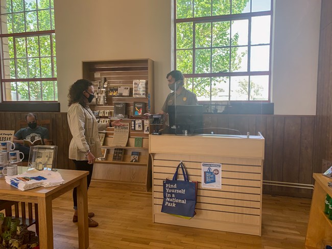 The Nicodemus Park Store with a man reading a book, and another costumer purchasing a postcard from a park ranger.