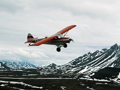 Small red airplane flying through mountains dotted with snow.