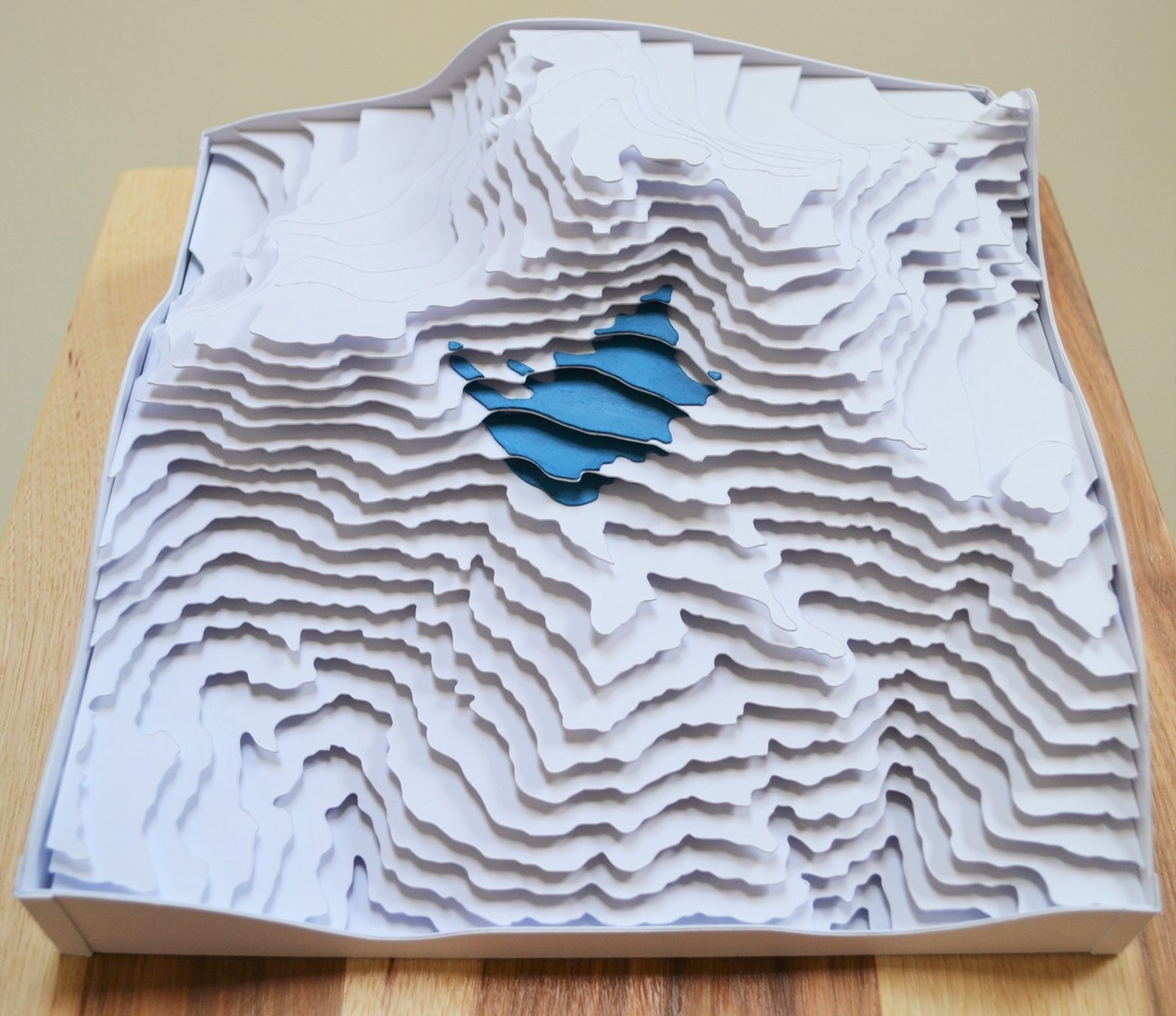 Layers of paper, cut and arranged in stacked horizontal planes to create the contours of a white mountain peak and a small blue glacier tucked just below the summit.