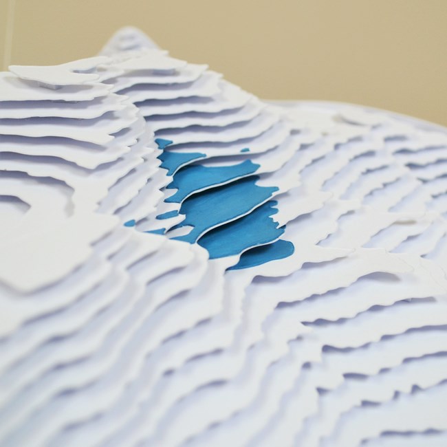 A closer view of layers of paper, cut and arranged in stacked horizontal planes to create the contours of a white mountain peak and a small blue glacier tucked just below the summit.