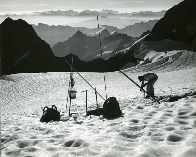 A man bends over equipment embedded in the ice of a huge glacier, including long polls, backpacks, and a bucket. In the distance, a panorama of mountains beyond mountains.