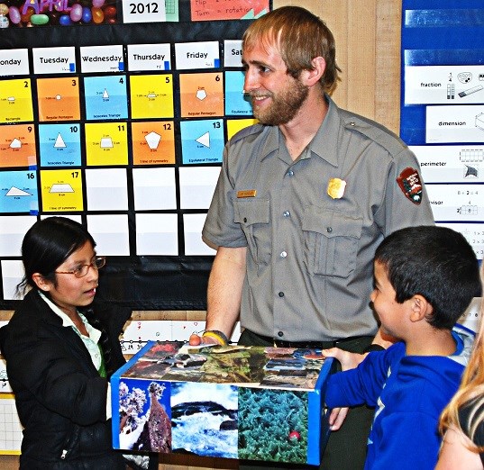 A ranger smiles as students reach into a box filled with natural items.