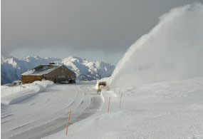 snowplow working in large snow-covered parking lot; mountains and building in background snowplow working in large snow-covered parking lot; mountains and building in background