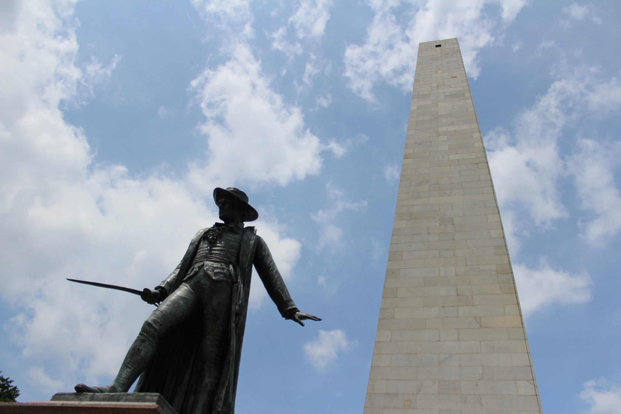 A statue of a man with a coat and hat on a cloudy day in front of a tall white-brick obelisk.