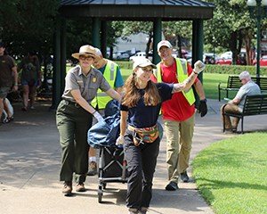 A woman holds a hand in the air while pulling a cart full of debris. A park ranger and two other people are pushing the cart.