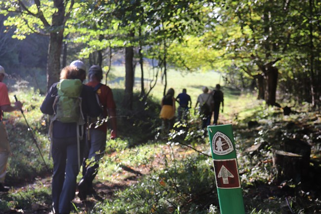 People hiking up a trail through a grassy pasture, passing a wooden fence post with a trail sign.