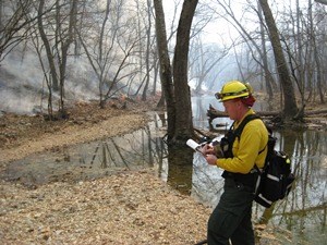 a firefighter checks notes next to a smoking line of fire in the woods