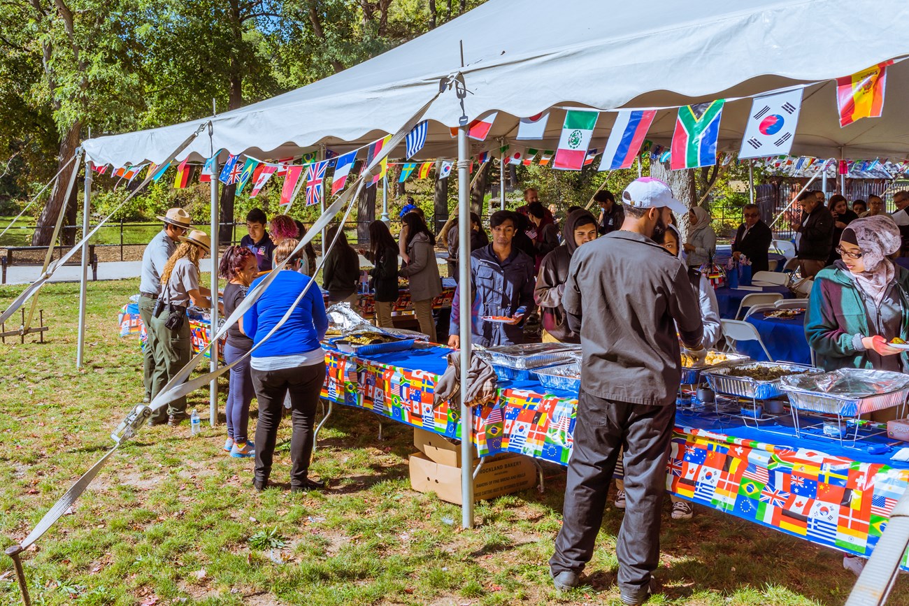 Park rangers & visitors fill plates of food at an outdoor multinational food event under a tent, flags of all nations hanging overhead