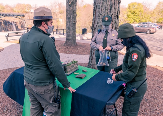 Three park rangers set up an outdoor activity table near a waterfall spanned by an arched bridge, wearing pandemic masks