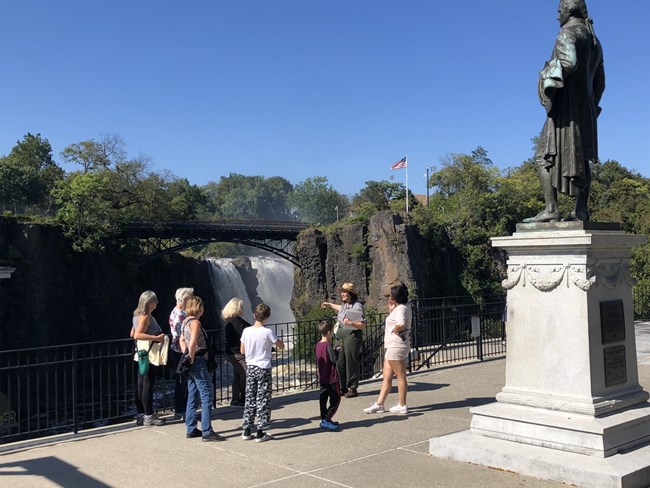 A park ranger leading a tour stands beside a statue of Alexander Hamilton, pointing behind her to a tall waterfall framed by dark basalt cliffs and an arched metal bridge. An American flag flies on a pole on the cliff above
