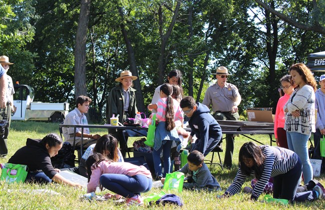 Children play in the grass surrounding park rangers at an outdoor activity table