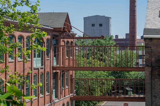 19th century brick mill buildings connected by metal walkways stand with green vegetation around them, tall factory structures & a chimney in the background