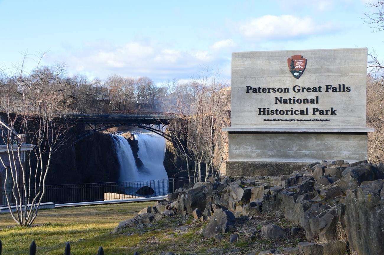A large sign for Paterson Great Falls National Historical Park, with the Great Falls in the background framed by an arched metal bridge