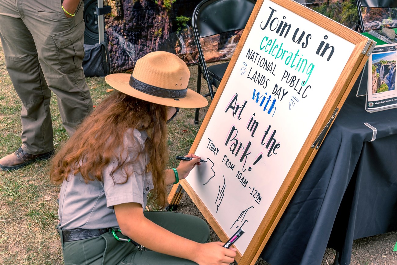 A park ranger writes information on an a-frame whiteboard about a National Public Lands day art in the park event at Paterson Great Falls National Historical Park