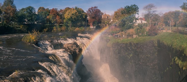 A rainbow forms over a roaring waterfall pouring into a deep chasm framed by trees, buildings, & an observation overlook with visitors