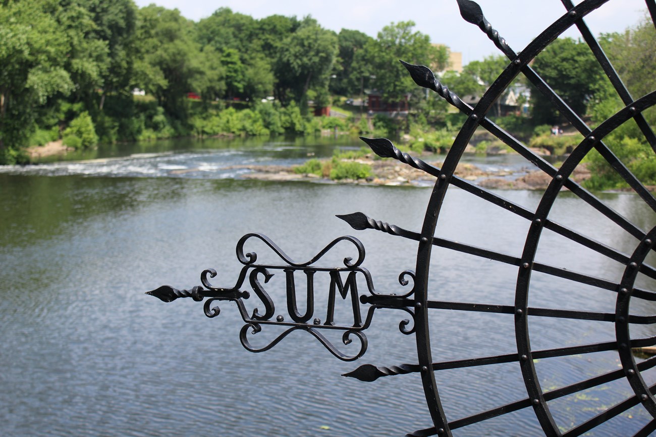 A half-moon black ornamental fence facing left with the letters "S.U.M." in ornate filigree. Facing left, it frames a view of the Passaic River & the upper edge of the Great Falls surrounded by trees