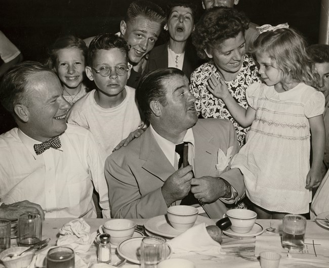 Black & white photo of Lou Costello at a table surrounded by smiling men, women, & children - he is turned, grinning, towards his young daughter, standing on a chair