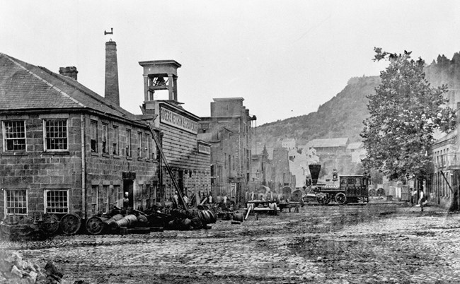 1850s black & white photo of a dirt street lined by stone, brick, & wood 2-3 story buildings, a mountain in the background. Closest at left is the Rogers works, an 0-4-0 steam locomotive, wheel castings, & cannons out front