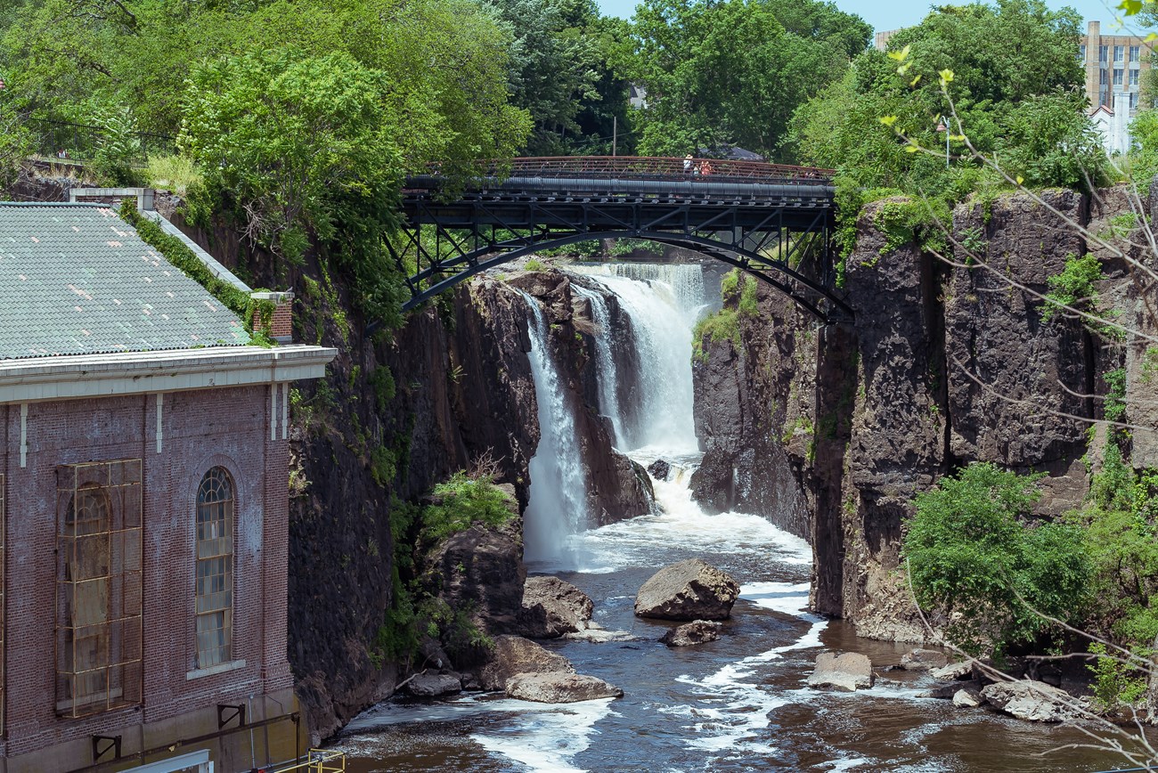 A 77 ft. waterfall plunging between dark basalt cliffs, framed by green vegetation and a black arched metal bridge. A brick building with tall windows is partially visible at left