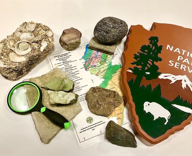A collection of rocks and minerals scattered amongst a National Park Service arrowhead, a magnifying glass, & a map of New Jersey's bedrock geology