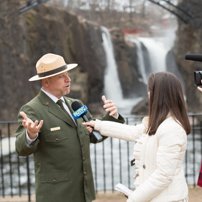 A park ranger in green dress uniform is interviewed by a news anchor in front of a roaring waterfall plunging between dark basalt cliffs below an arched black metal bridge