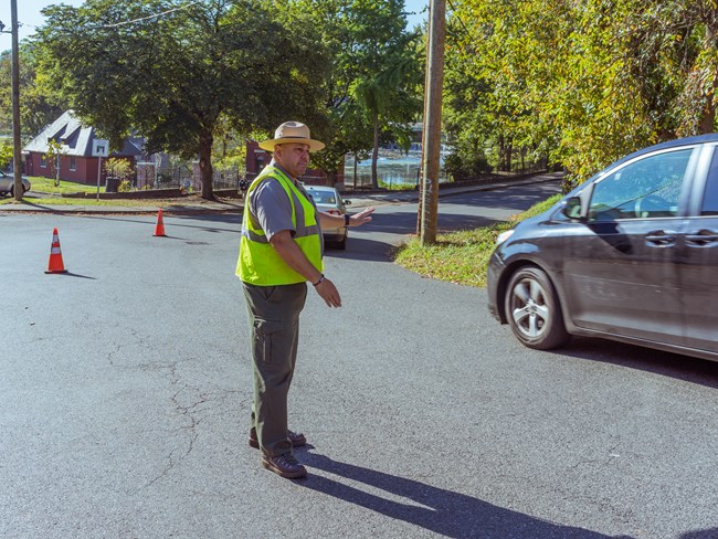 A park ranger with a fluorescent safety vest directs cars past traffic cones on a street line with trees, brick buildings visible in the park behind