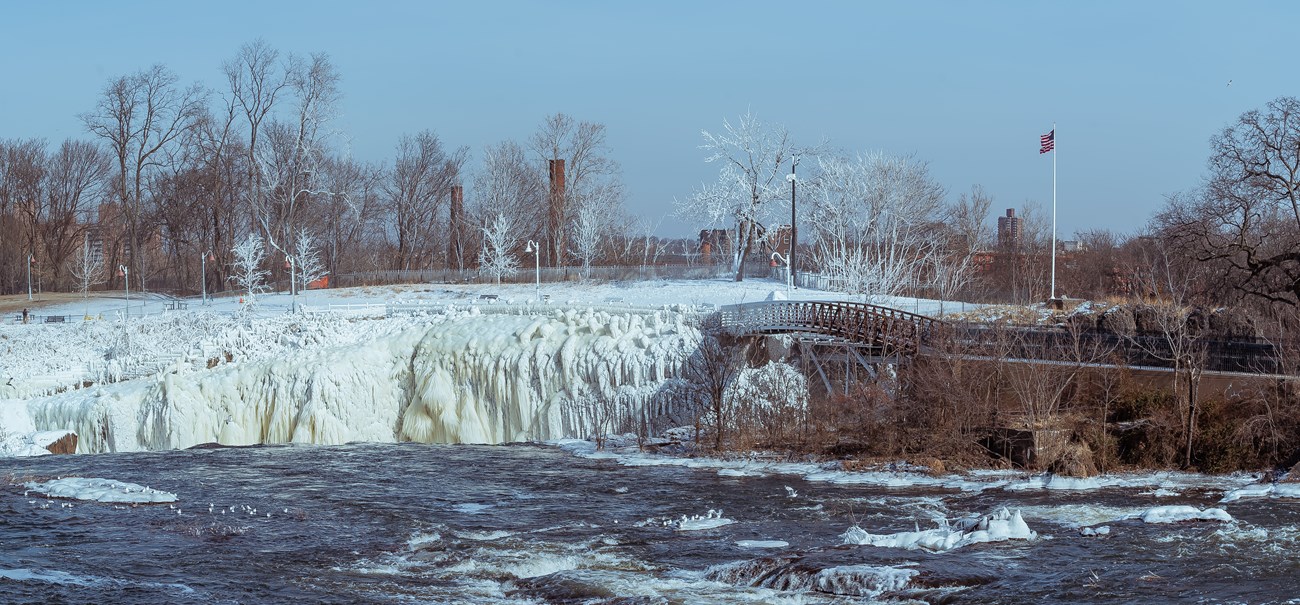 A wide view of a waterfall pouring into a chasm spanned by a bridge at right, an American flag high on a pole. The area around the falls & closest side of the bridge are covered in thick coatings of heavy white ice