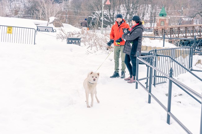 Visitors in black & orange winter jackets stand in deep snow, area barricades, benches, & trash cans partially buried. One holds a small dog with a sweater in her arms - the other has a large white dog on a leash