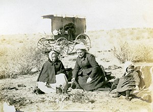 Two women and a baby picnic on the ground in front of a wagon