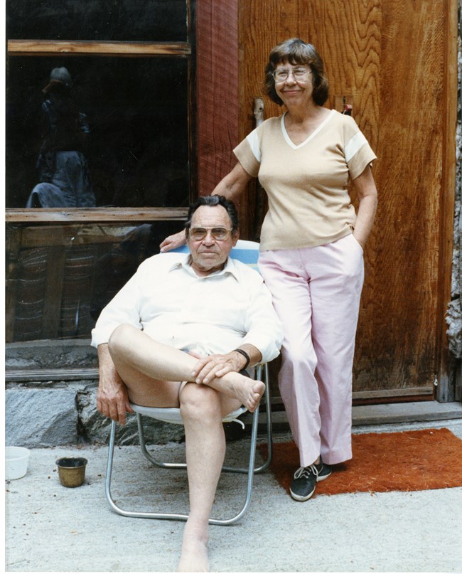A woman in her sixties stands behind a man, seated, in his sixties at the front entryway of a house.