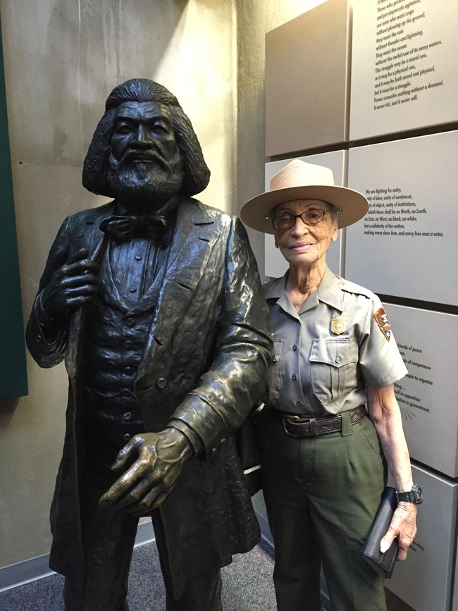 An image of Betty Reid Soskin posing for a picture next to a statue to her right.