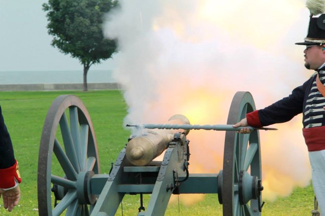 Reenactor stands on the right side of cannon on a field carriage and lights the cannon. Smoke & fire come out the muzzle of the cannon.