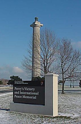 Tall memorial column with a sign in front that says Perry's Victory and International Peace Memorial. There is snow on the ground a couple of trees without leaves in the background.