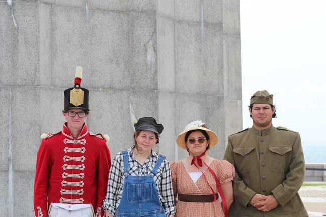 Four people wearing historic clothing. Left is a British War of 1812 infantryman. Next is a person dressed in overalls and blue and white flannel shirt, Then a person wearing a reddish/brown floral print regency dress. The right most is wearing a US olive
