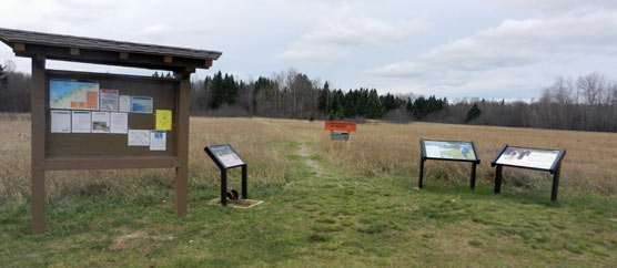 Trailhead at the Grand Sable Visitor Center with its bulletin board, boot brush station & wayside exhibit, trail, backcountry regulations & mileage sign, and set of two North Country National Scenic Trail wayside exhibits.