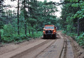 An orange dump truck with grader works on the gravel surface of Alger County Road H-58.