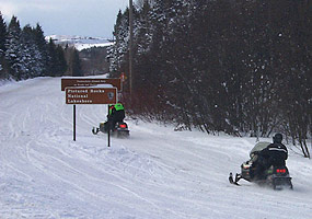 Winter visitors operating snowmobiles travel along County Road H-58 with the Grand Sable Dunes rising in the background.