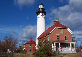 The red brick double keepers quarters and white light tower of the Au Sable Light Station stand proud before the blue sky.