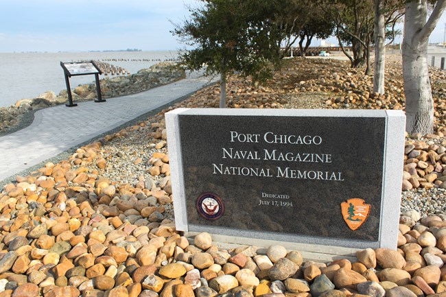 A granite sign reads "Port Chicago Naval Magazine National Memorial" and sits in front of trees and stones. A sidewalk can be seen on the left side.