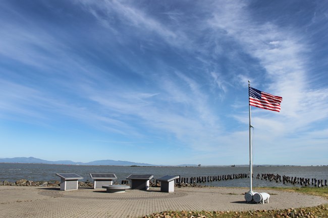 Four memorial granite stones are arranged in an arc. The bay water and hills can be seen in the background. An American flag on a a pole stands next to the memorial stands.
