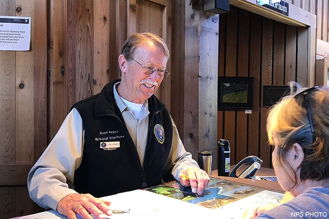 A smiling man dressed in a National Park Service volunteer uniform stands behind a desk on with maps displayed while talking with a visitor.